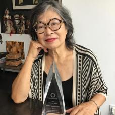 Female with black and grey hair and tortoiseshell round glasses is behind a table holding her Visionary glass trophey